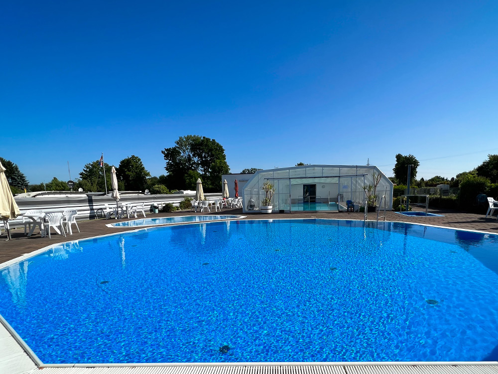 Rundes Schwimmbad im Freien, umgeben von weißen Stühlen und Sonnenschirmen, mit einem gläsernen Gebäude und Bäumen im Hintergrund unter einem klaren blauen Himmel.