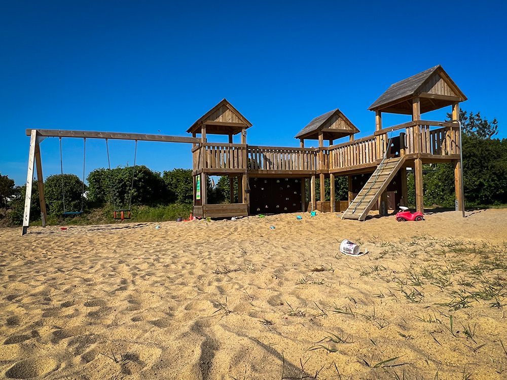 Ein hölzerner Spielplatz mit Rutschen, Brücken und Schaukeln steht auf einer sandigen Fläche unter einem strahlend blauen Himmel; einige verstreute Spielsachen sind im Sand zu sehen.