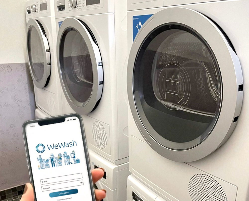 Eine Person hält ein Smartphone mit der WeWash-App vor die gestapelten Waschmaschinen in einer Waschküche.