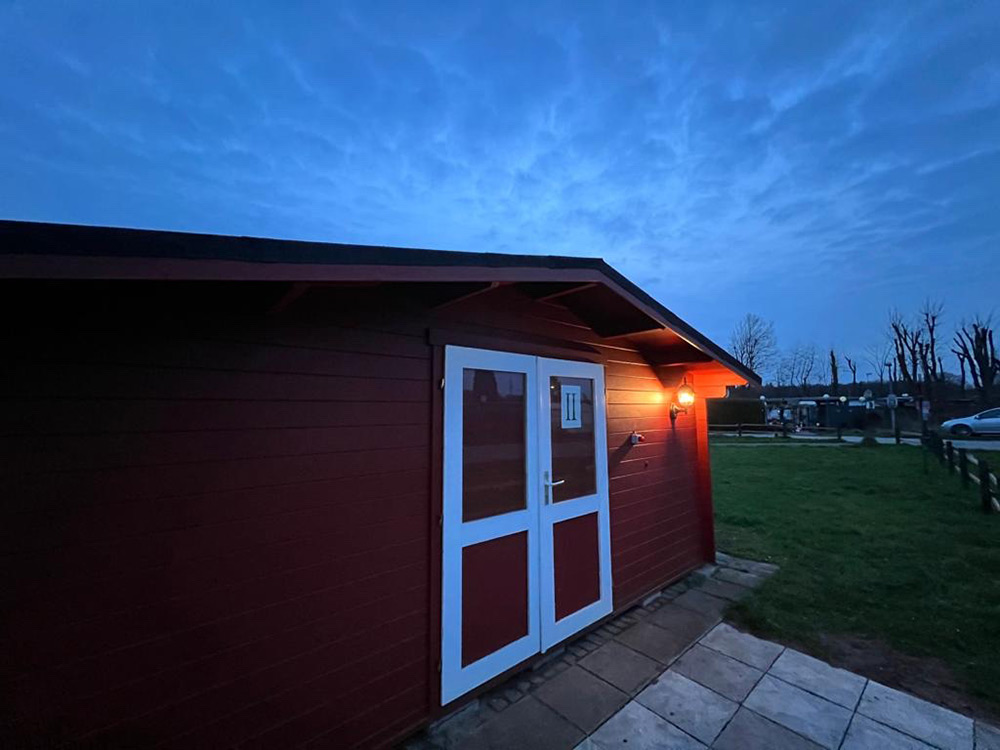 Ein kleines rotes Holzgebäude mit weißen Flügeltüren, das von einer Außenwandlampe beleuchtet wird, steht in der Abenddämmerung unter bewölktem Himmel auf einer Wiese.