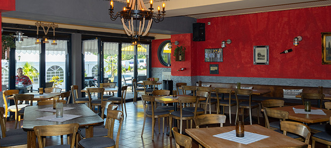 Ein Restaurant mit Holztischen und -stühlen, roten und grauen Wänden, gerahmten Bildern und großen Fenstern, die natürliches Licht hereinlassen.