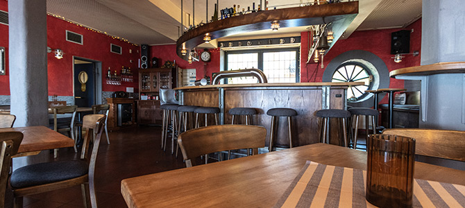Eine gemütliche Bar mit Holztischen und -hockern, roten Wänden und einem geschwungenen Tresen, durch dessen Fenster natürliches Licht fällt.