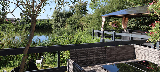 Die Außenterrasse mit Korbmöbeln und einem Pavillon bietet an sonnigen Tagen einen Blick auf einen Teich, der von grünen Bäumen und Sträuchern umgeben ist.
