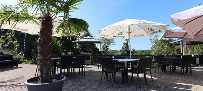 Eine Terrasse im Freien mit schwarzen Tischen und Stühlen, großen Sonnenschirmen als Schattenspender, einer getopften Palme und Grünpflanzen im Hintergrund unter einem klaren blauen Himmel.