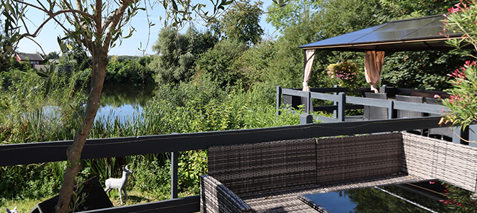 An einem sonnigen Tag überblicken eine Korbveranda und ein Pavillon einen Teich, der von üppigem Grün und Bäumen umgeben ist.