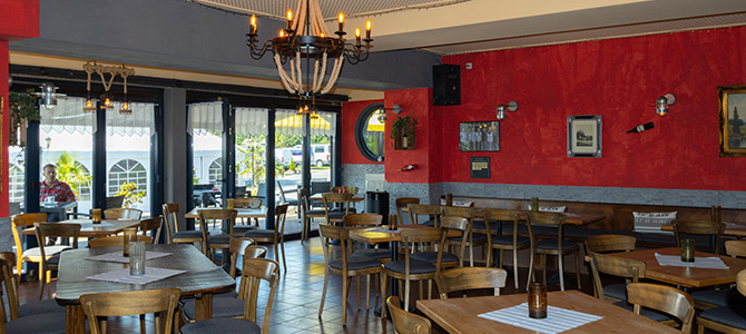 Ein Restaurant mit Holztischen und -stühlen, roten und grauen Wänden, Kronleuchtern und großen Fenstern, die natürliches Licht hereinlassen.