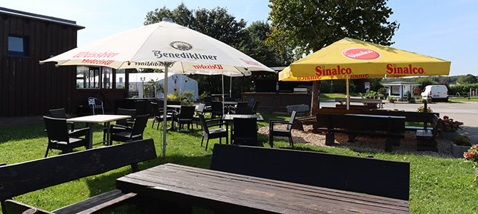 Sitzgelegenheiten im Freien mit Holzbänken und -tischen, die an sonnigen Tagen von großen Sonnenschirmen mit den Logos von Benediktiner und Sinalco beschattet werden.