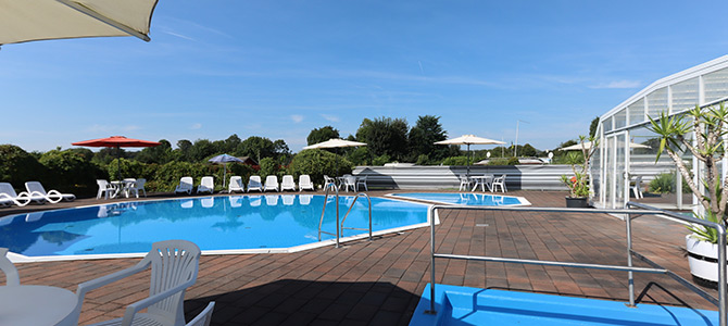 Außenschwimmbad, umgeben von Liegestühlen, Sonnenschirmen und Grünpflanzen unter blauem Himmel, mit einer Glaskonstruktion auf der rechten Seite.